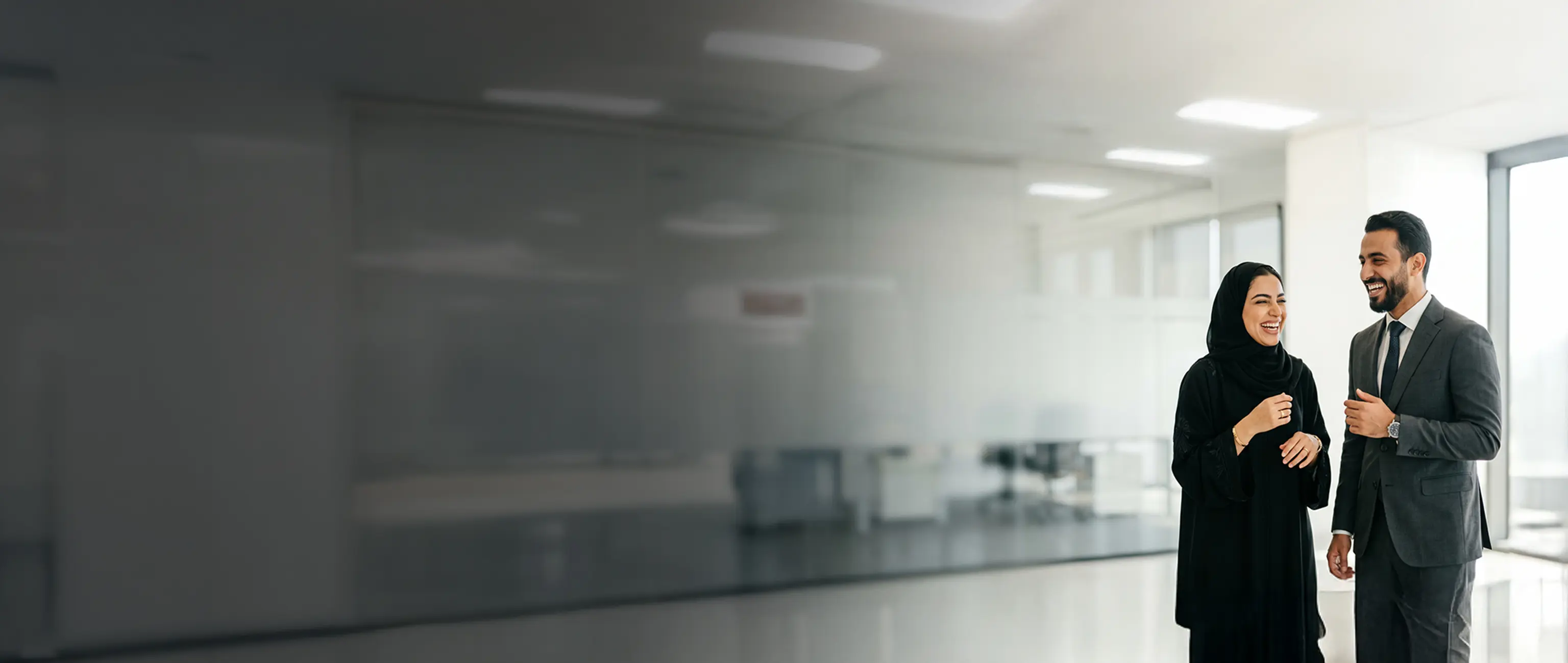 A businessman in a formal suit and a woman in an abaya talking and smiling in a modern corporate workplace, representing the Employee Savings Plan from National Bonds.