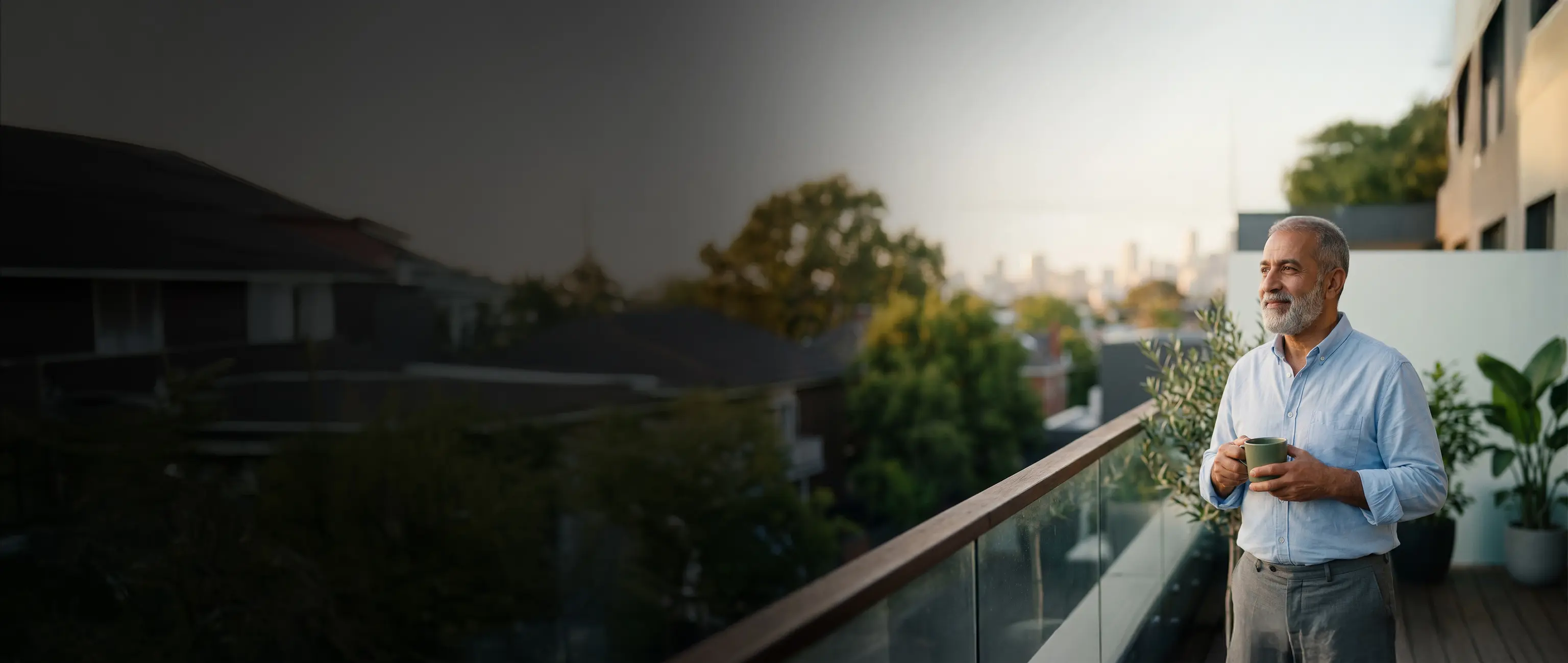 An older man holding a mug and looking out peacefully from a balcony at a scenic landscape and city skyline, representing comfort and financial security for retirement with the End of Service program from National Bonds.