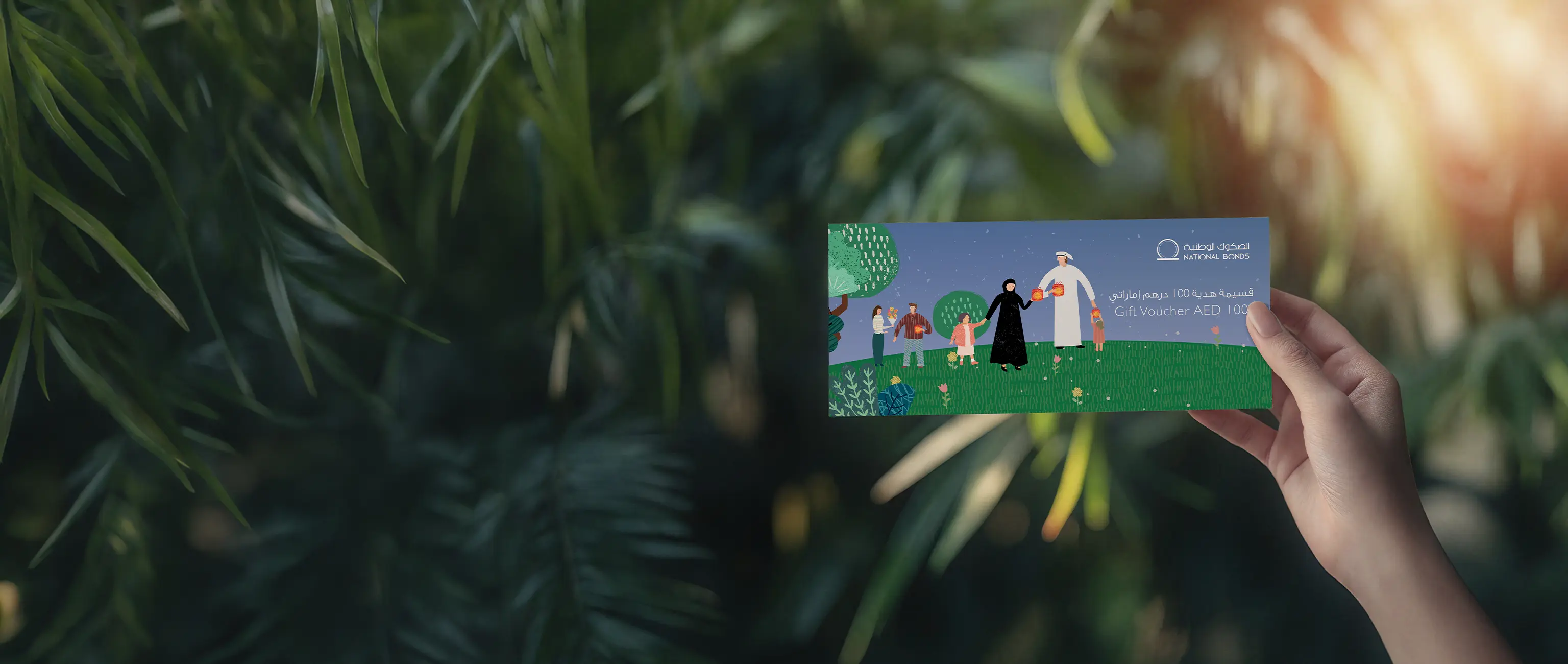 A hand holding an AED 100 National Bonds gift voucher featuring an illustration of families, against a natural green foliage background, representing the "Gift Vouchers" product to encourage the culture of saving and gifting a bright financial future.